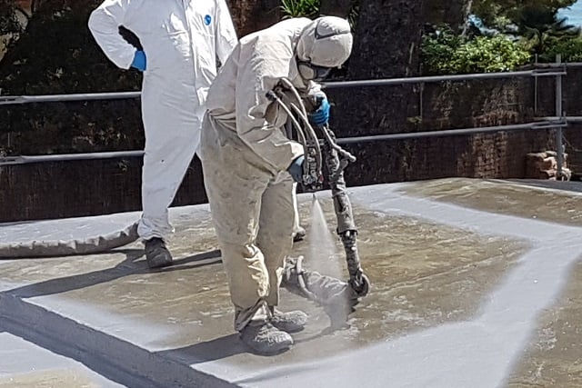 Técnicos operarios vestidos con equipos de protección, mono de trabajo, casco y máscara de protección proyectando espuma de poliuretano en una cubierta exterior