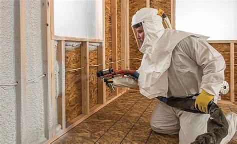 Técnico operario vestido con equipo de protección, mono de trabajo, casco y máscara de protección proyectando espuma de poliuretano en el interior de una cámara a través de un orificio en la pared